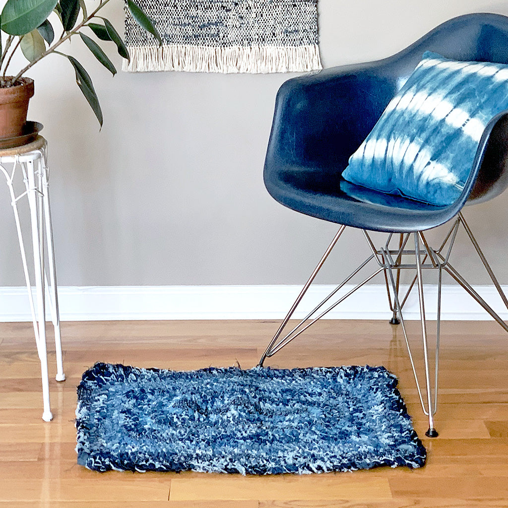 Hand Woven Upcycled Denim &quot;Amish Knot&quot; Rug. Rectangular shaped with different rows of varied coloured indigo on a wooden floor with blue chair and plant on a plant stand. 